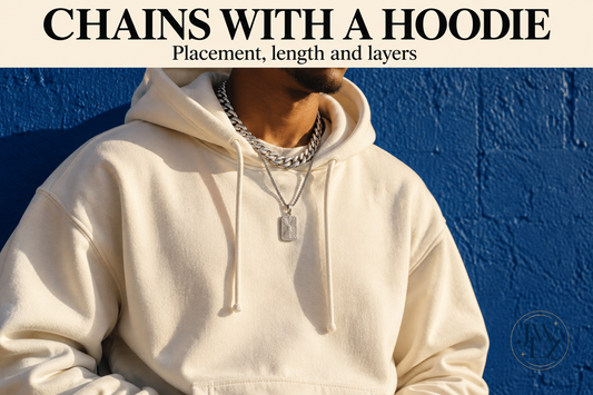 Editorial concept of a short Cuban-style chain and a separate lower pendant chain on a cream hoodie against a cobalt wall.